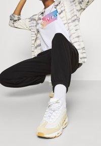 Vit t-shirt med flerfärgad Levi's-logga, svarta joggers med elastiska muddar och vita/gula sneakers med texturerade accenter.