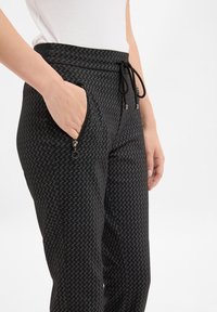 Schwarze gemusterte Jogginghose mit einem Kordelzugbund, seitlichen Zippertaschen und einem weichen Stoffgefühl. Das Zickzackmuster ist subtil und gleichmäßig.