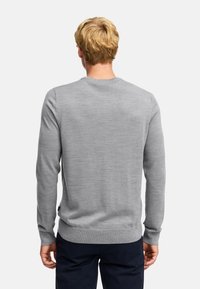 Uomo con capelli biondi che indossa un maglione a maniche lunghe grigio chiaro semplice e pantaloni scuri, girato di spalle su uno sfondo bianco.