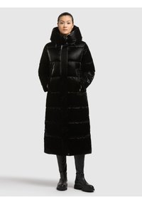 Garš melns pūkains mētelis ar polsterētu dizainu, lielu kapuci un priekšējo pogu aizdari. Aprīkots ar sānu kabatām un gludu, spīdīgu faktūru.