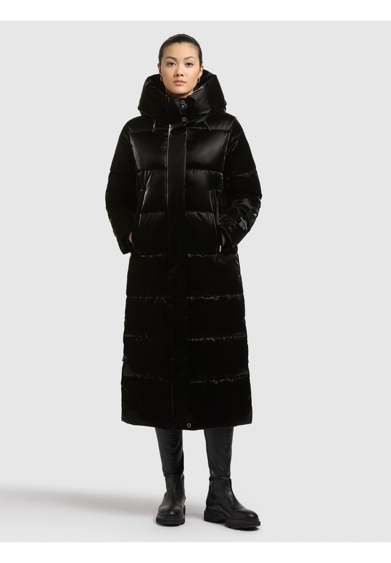 Garš melns pūkains mētelis ar polsterētu dizainu, lielu kapuci un priekšējo pogu aizdari. Aprīkots ar sānu kabatām un gludu, spīdīgu faktūru.