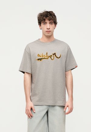 Νέος άνδρας με σγουρά καστανά μαλλιά, φορώντας γκρι φαρδιά μπλούζα με πορτοκαλί γραφικό κείμενο και ανοιχτό μπλε φαρδιά τζιν, στέκεται μπροστά σε λευκό φόντο.