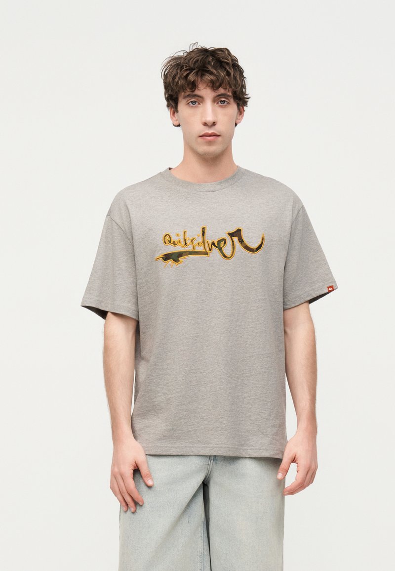 Νέος άνδρας με σγουρά καστανά μαλλιά, φορώντας γκρι φαρδιά μπλούζα με πορτοκαλί γραφικό κείμενο και ανοιχτό μπλε φαρδιά τζιν, στέκεται μπροστά σε λευκό φόντο.