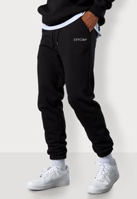Schwarze Baumwoll-Sweatpants mit elastischen Bündchen, mit einem weißen Logo "ESTEEM+" im linken Taschenbereich. Kombiniert mit weißen Sneakers.