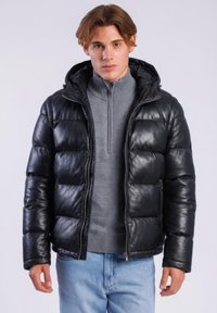 Jonge man draagt een zwarte donsjas met capuchon, een grijze ritsvest en lichtblauwe spijkerbroek, staand tegen een lichtgrijze achtergrond.