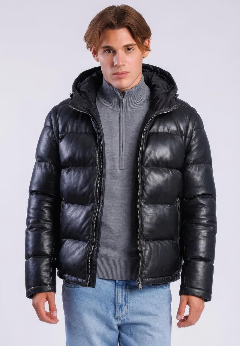 Jonge man draagt een zwarte donsjas met capuchon, een grijze ritsvest en lichtblauwe spijkerbroek, staand tegen een lichtgrijze achtergrond.