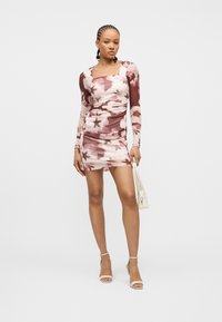 Γυναίκα με πλεγμένα μαλλιά, που φοράει ένα ροζ και λευκό μίνι φόρεμα με μακριά μανίκια και λουλουδάτο σχέδιο, κρατώντας μια μικρή λευκή τσάντα και φορώντας λευκά τακούνια με λουράκι στον αστράγαλο.