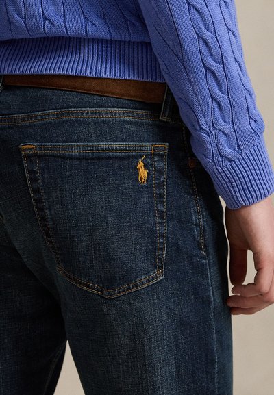 Άτομο που φοράει σκούρο μπλε τζιν με κίτρινο κεντημένο λογότυπο στην πίσω τσέπη και μπλε πουλόβερ πλεκτό με καλώδια και καφέ ζώνη.