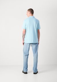 Lyseblå polo skjorte med korte ærmer, lavet af glat stof. Matchet med lyse denim jeans og sorte sko, set fra ryggen.