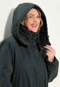 Chaqueta impermeable verde oscuro con capucha, que presenta un alto cuello, cierres de botón y puños elásticos para mayor comodidad. Forro interior negro visible.
