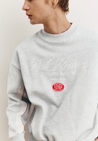 Grauer Pullover mit geprägtem "Pull Records"-Text und einem roten, runden Aufnäher mit der Aufschrift "DEPARTMENT SPORTS P&BR" auf der Brust.