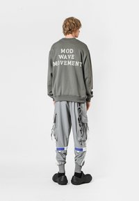 Sudadera gris con "MOVIMIENTO MOD WAVE" impreso en la espalda, combinada con pantalones cargo grises holgados que cuentan con múltiples bolsillos y zapatos negros sin cordones.