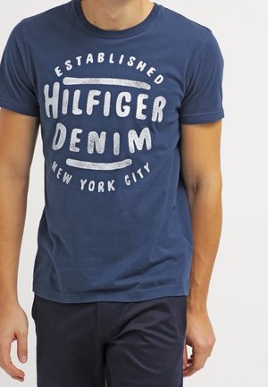 Mężczyzna ubrany w granatowy T-shirt z białym napisem "Established Hilfiger Denim New York City" oraz ciemne spodnie, stojący na jednolitym tle.