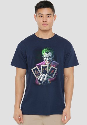 Young man wearing a navy shirt with a graphic of the Joker holding four playing cards, standing against a plain background.