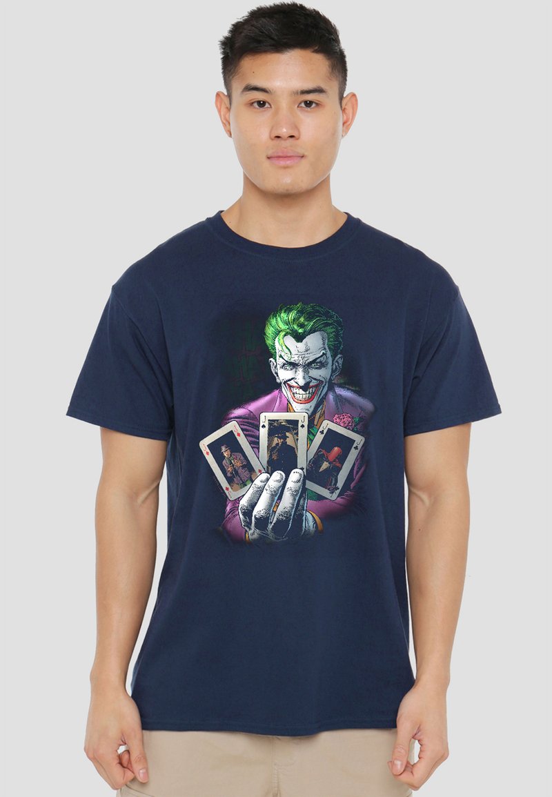 Young man wearing a navy shirt with a graphic of the Joker holding four playing cards, standing against a plain background.