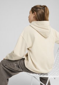 Sudadera de gran tamaño en color crema con una textura suave y una capucha grande, combinada con pantalones de mezclilla gris, frente a un fondo neutro.