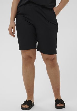 Személy fekete térdig érő rövidnadrágot és fekete papucsot visel, egyik kezével a zsebében áll, egyszínű háttér előtt.