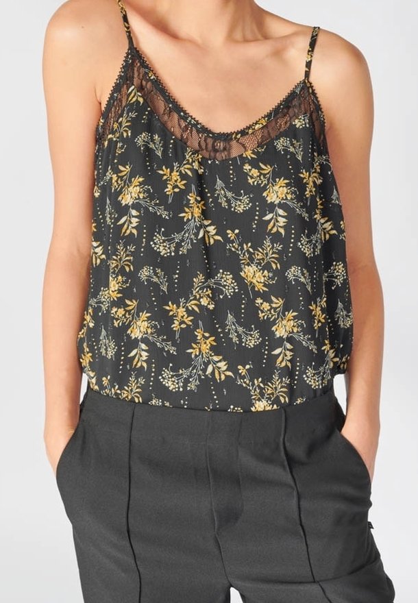 Camisole noire à motif floral avec bordure en dentelle associée à un pantalon taille haute foncé, mains dans les poches, montrée des épaules aux hanches.