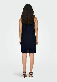 Robe sans manches bleu marine avec un ourlet en dentelle texturée, présentant des motifs complexes. Tissu doux, longueur genou, associée à des sandales à talons noirs.