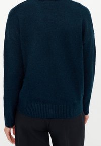 Mörkt tealstickad tröja med avslappnad passform, ribbat nederkant och manschetter. Baksidan har minimala detaljer med en mjuk, texturerad yta.