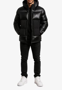 Doudoune noire avec capuche, dotée d'une fermeture éclair et d'un logo. Associée à un jean noir et des baskets blanches. Texture lisse et brillante.