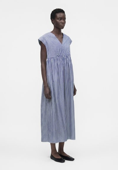 Γυναίκα στέκεται φορώντας ένα midi φόρεμα χωρίς μανίκια με κάθετες μπλε και άσπρες ρίγες και μαύρα επίπεδα παπούτσια σε απλή φόντο.
