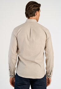 Uomo che indossa una camicia beige a maniche lunghe con bottoni e pantaloni scuri, visto di spalle su uno sfondo chiaro e semplice.
