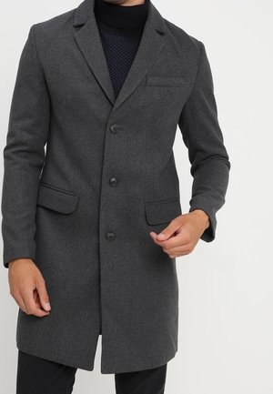 Homme portant un manteau en laine gris foncé boutonné par-dessus un pull à col roulé noir, debout devant un fond clair uni.