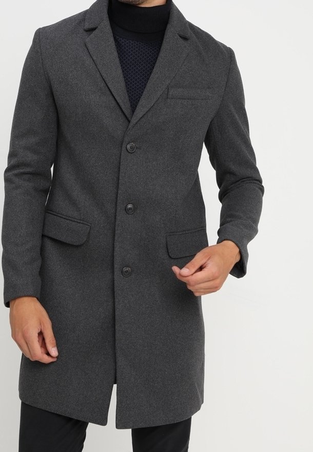 Homme portant un manteau en laine gris foncé boutonné par-dessus un pull à col roulé noir, debout devant un fond clair uni.