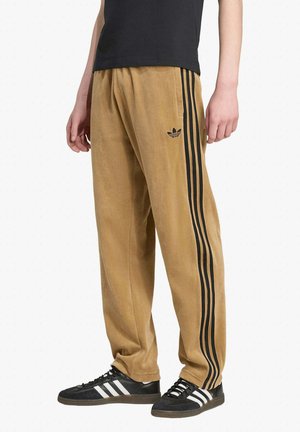 Persona che indossa pantaloni sportivi Adidas color cuoio con triple strisce nere e scarpe da ginnastica Adidas nere con strisce bianche, in piedi davanti a uno sfondo bianco.