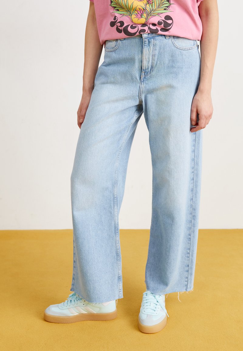 Jeans alta vita di colore azzurro chiaro con taglio a gamba larga e orlo grezzo, abbinati a sneaker verde menta. La t-shirt presenta un vivace design floreale.