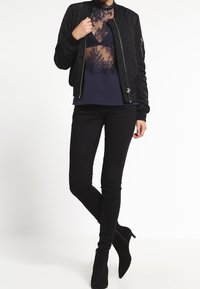 Giubbotto bomber nero con polsini a coste, indossato sopra una maglietta trasparente blu navy con dettagli in pizzo, abbinato a jeans skinny neri e stivaletti.
