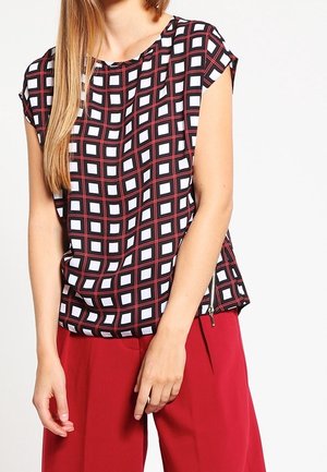 Kobieta ubrana w czarno-biało-czerwoną kratowaną bluzkę z krótkim rękawem w połączeniu z czerwonymi, szerokimi spodniami z wysokim stanem na jednolitym tle.