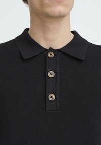 Černé polo tričko s texturovanou pleteninou, vybavené límcem a čtyřmi světlými knoflíky na légu.