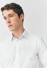 Weißes Hemd mit klassischem Kragen, Knopfleiste und glatter Textur. Verfügt über eine einzelne Brusttasche und eine taillierte Passform.