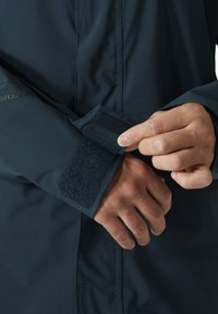 Water-resistant jacket with a dark blue material. Features a Velcro cuff adjustment and subtle branding on the sleeve. Smooth texture.