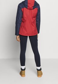 Röd och marinblå vattentät jacka med huva, matchad med marinblå leggings och tanfärgade ankelstövlar. Slät yta och atletisk passform.