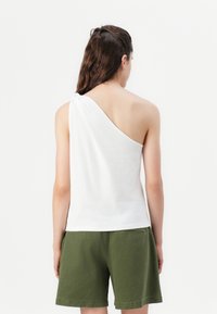 Top alb cu un umăr, realizat dintr-un material moale, având un detaliu legat pe umăr. Asortat cu pantaloni scurți verzi, cu un design pronunțat la spate.