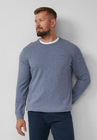 Mann mit Bart, der einen blauen Rundhalsausschnitt-Pullover über einem weißen Hemd und dunklen Hosen trägt, steht vor einer schlichten, hellen Wand und schaut nach links.