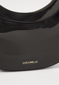 Svart lakkert lærveske med en buet silhuett, med en gull "Coccinelle"-logo nederst og tonale sømdetaljer.