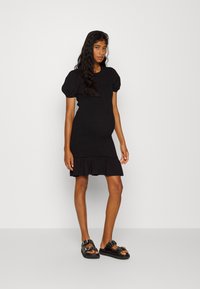 Vestido preto de malha com mangas bufantes, corpete ajustado e bainha franzida. Combinado com sandálias pretas robustas com tachas prateadas e tiras.