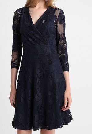 Robe en dentelle bleu marine avec manches trois-quarts, décolleté en V, motif floral, corsage ajusté et jupe évasée. Détail de tissu texturé sur toute la pièce.