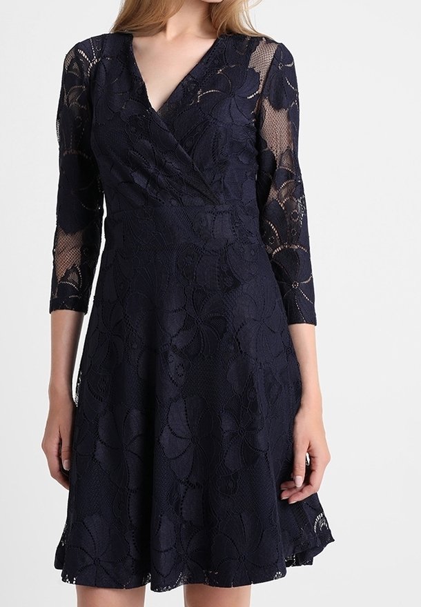 Robe en dentelle bleu marine avec manches trois-quarts, décolleté en V, motif floral, corsage ajusté et jupe évasée. Détail de tissu texturé sur toute la pièce.