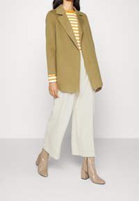 Blazer verde oliva, camicia a maniche lunghe bianca e arancione a righe, pantaloni larghi color avorio e stivaletti beige con tacco quadrato.