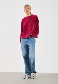 Pull en tricot rouge texturé avec un col rond, associé à un jean en denim bleu clair. Le modèle porte des baskets beiges et teal. Fond uni.