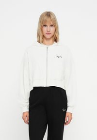 Gekürzte weiße Kapuzenjacke mit Reißverschluss, die über eine Rippstruktur und Logodetails verfügt. Getragen über schwarzen Ripp-Hosen mit passendem Logo.