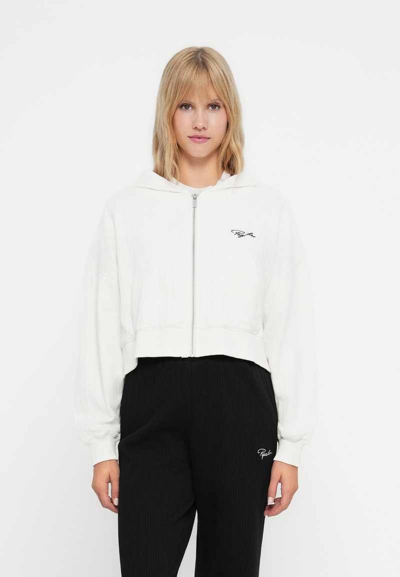 Gekürzte weiße Kapuzenjacke mit Reißverschluss, die über eine Rippstruktur und Logodetails verfügt. Getragen über schwarzen Ripp-Hosen mit passendem Logo.