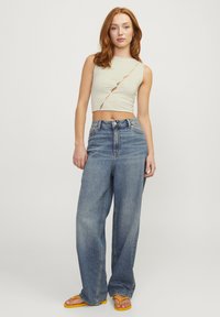 Top corto verde chiaro con ritagli diagonali, abbinato a jeans blu a vita alta e larghi, e infradito gialle.