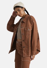 Giacca di jeans oversize marrone con bottoni neri, cuciture bianche a contrasto e due tasche frontali. Abbinata a una maglietta beige e pantaloni coordinati.