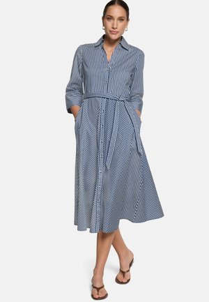 Woman wearing a blue and white striped button-down midi dress with 3/4 sleeves, a belt at the waist, with hands in pockets and brown sandals.